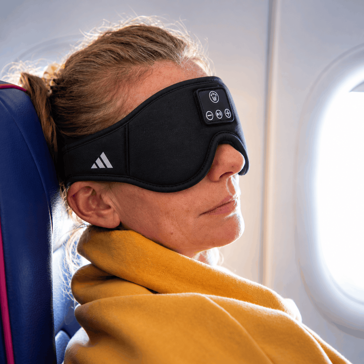 Black Wellable Bluetooth sleep mask with built-in audio controls and a white Adidas logo, worn by a woman resting in an airplane seat beside a window.