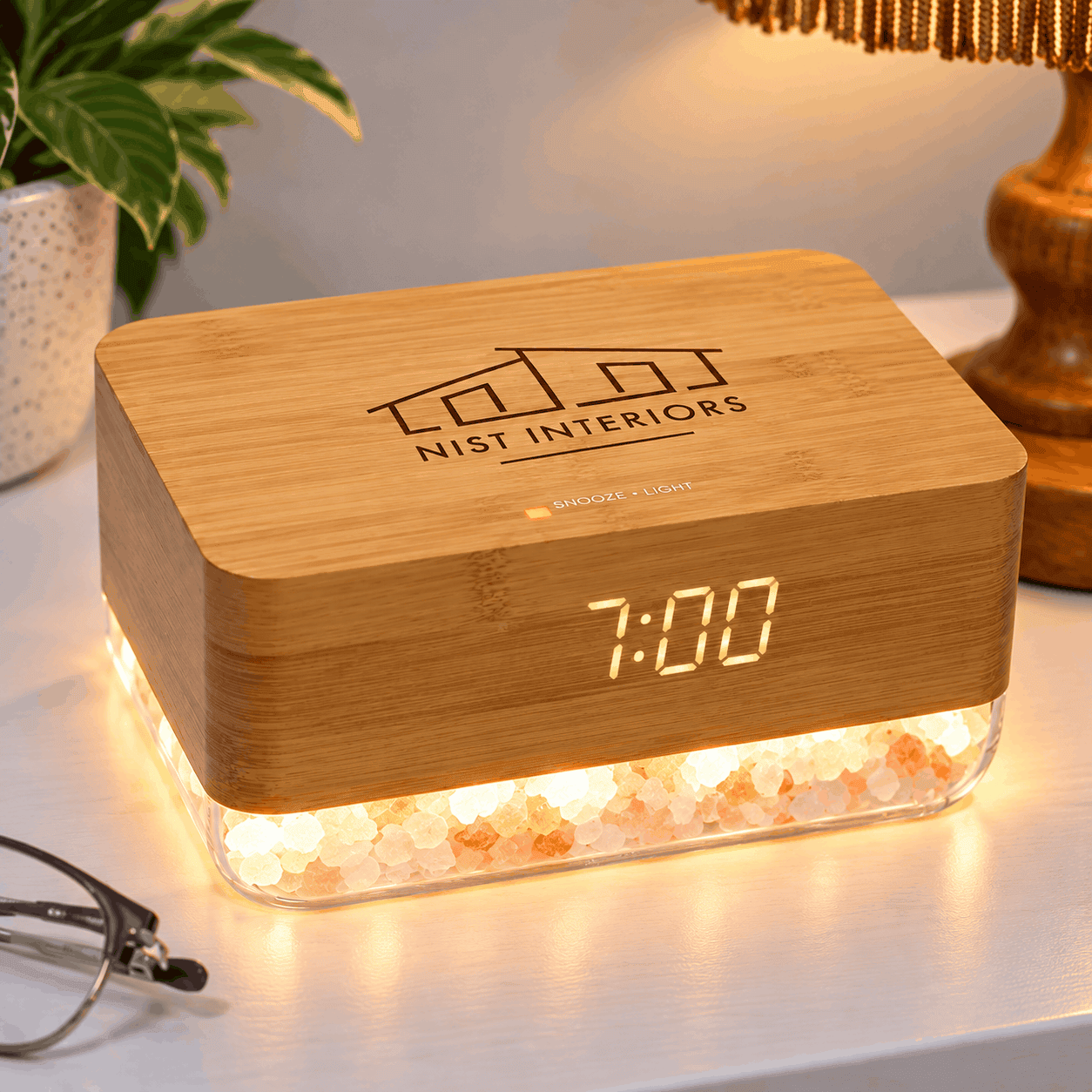 Bamboo sunrise alarm clock with a custom NIST Interiors logo, illuminated digital display, and glowing Himalayan salt crystals, sitting on a bedside table.
