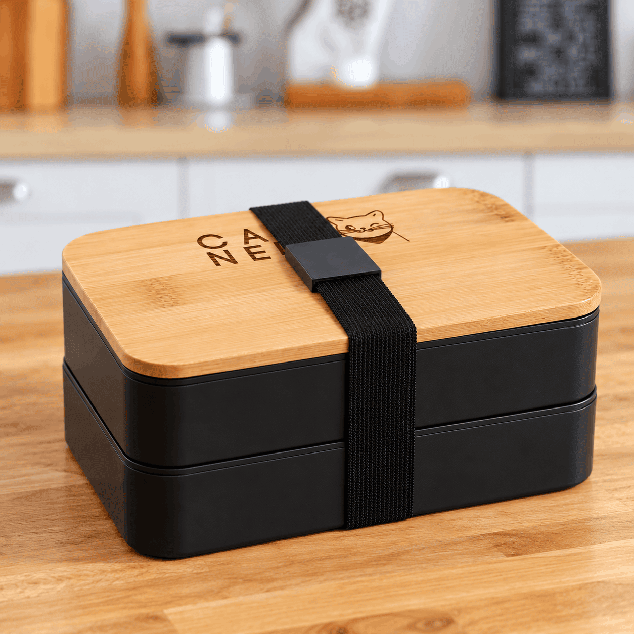 Black two-tier stackable bento box with a natural bamboo lid, custom engraved logo, and black elastic strap, displayed on a wooden kitchen countertop.