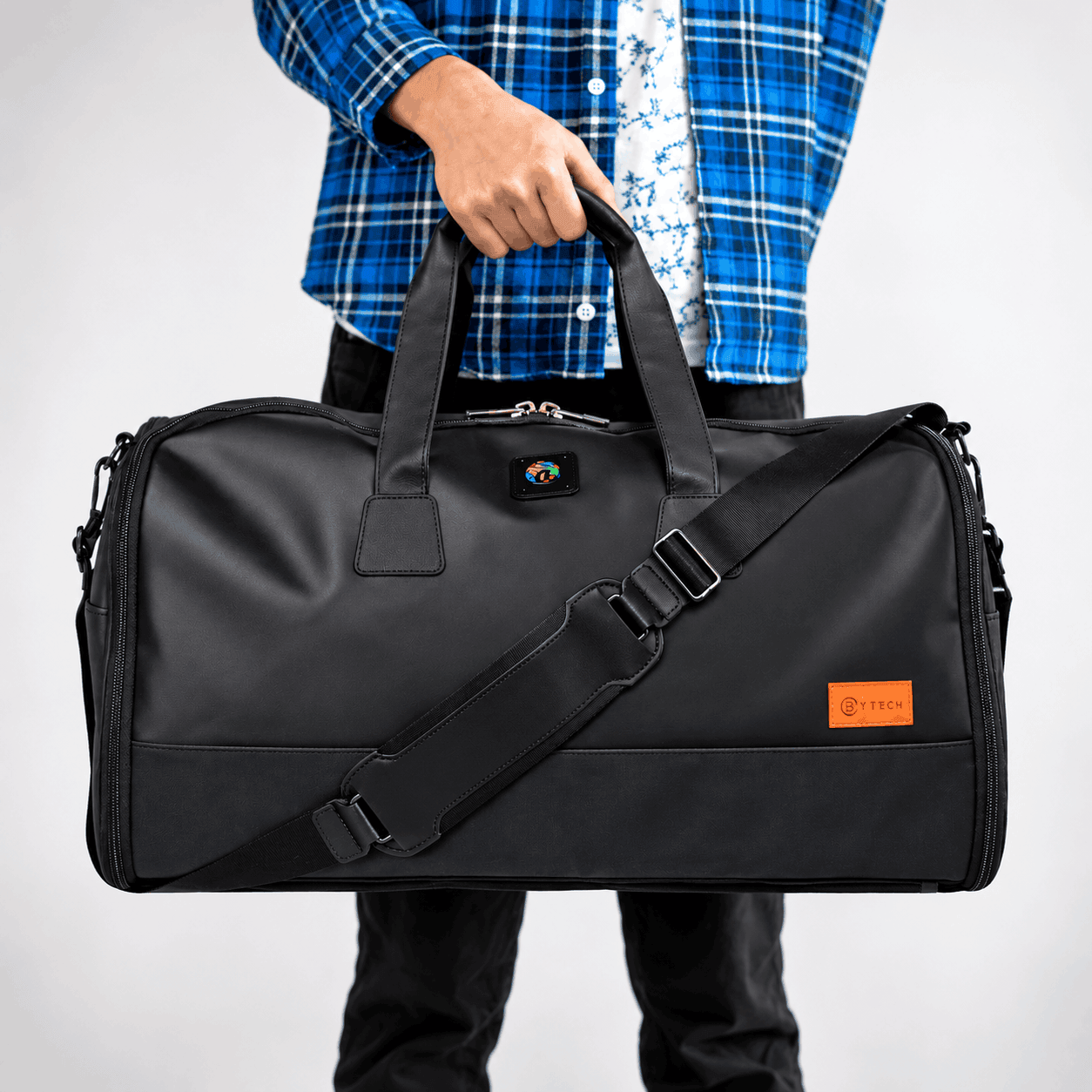 Black STITCH Ultimate Garment Bag with a custom embroidered logo, orange accent label, dual carrying handles, and adjustable shoulder strap, held by a man in a blue plaid shirt.