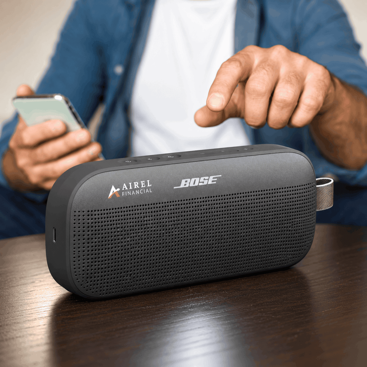 Black Bose SoundLink Flex 2 Bluetooth speaker with a custom Airel Financial logo, textured front grille, and carrying loop, displayed on a wooden table beside a person using a smartphone.