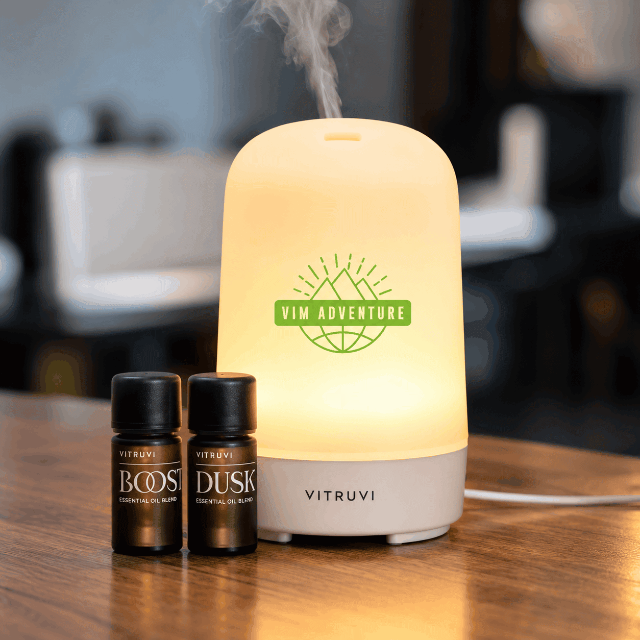 Glowing Vitruvi essential oil diffuser with a green Vim Adventure logo, releasing a gentle mist beside two essential oil bottles on a wooden tabletop.