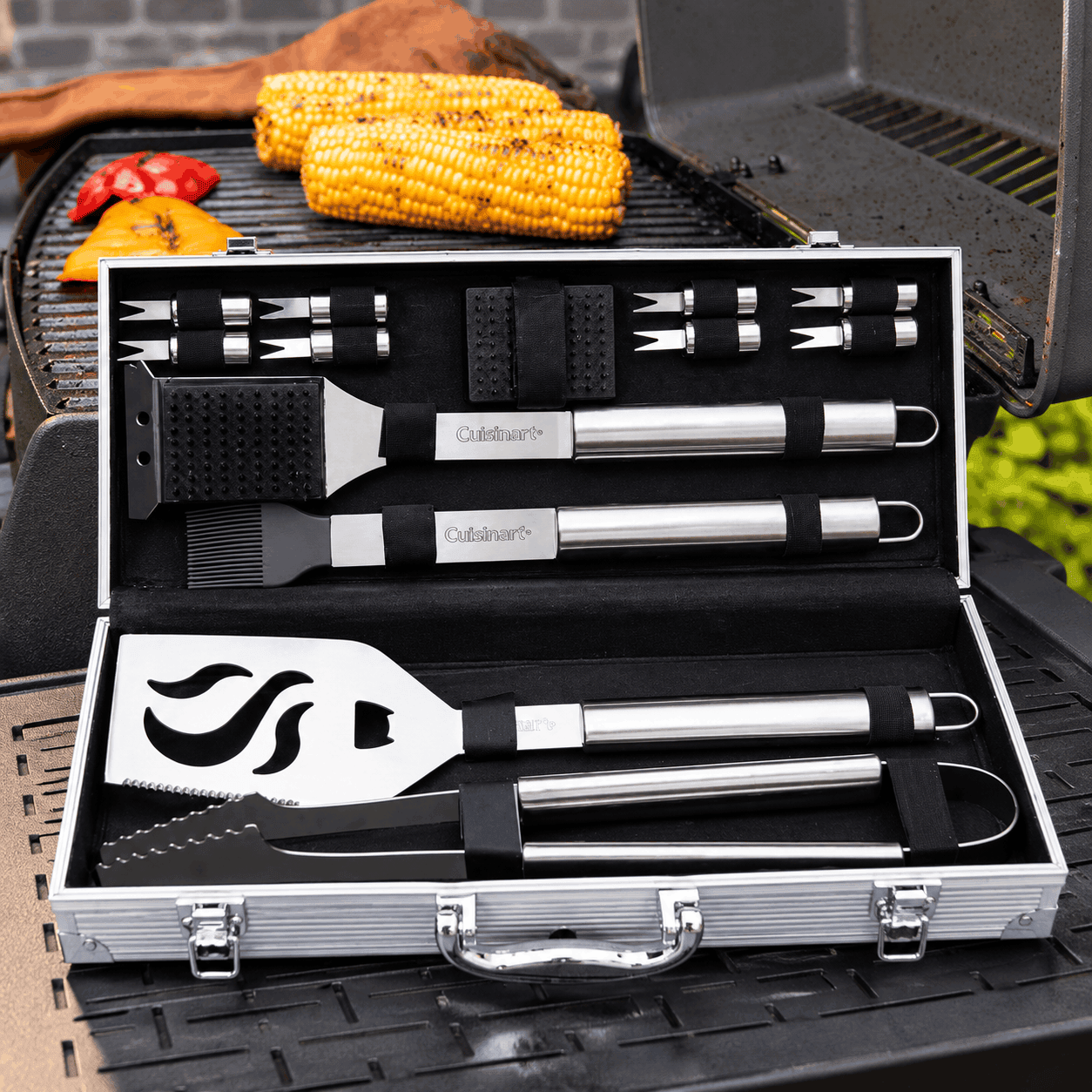 Cuisinart Outdoors 14-piece deluxe stainless steel grill tool set displayed in an open aluminum carrying case beside a barbecue grill with corn and vegetables.