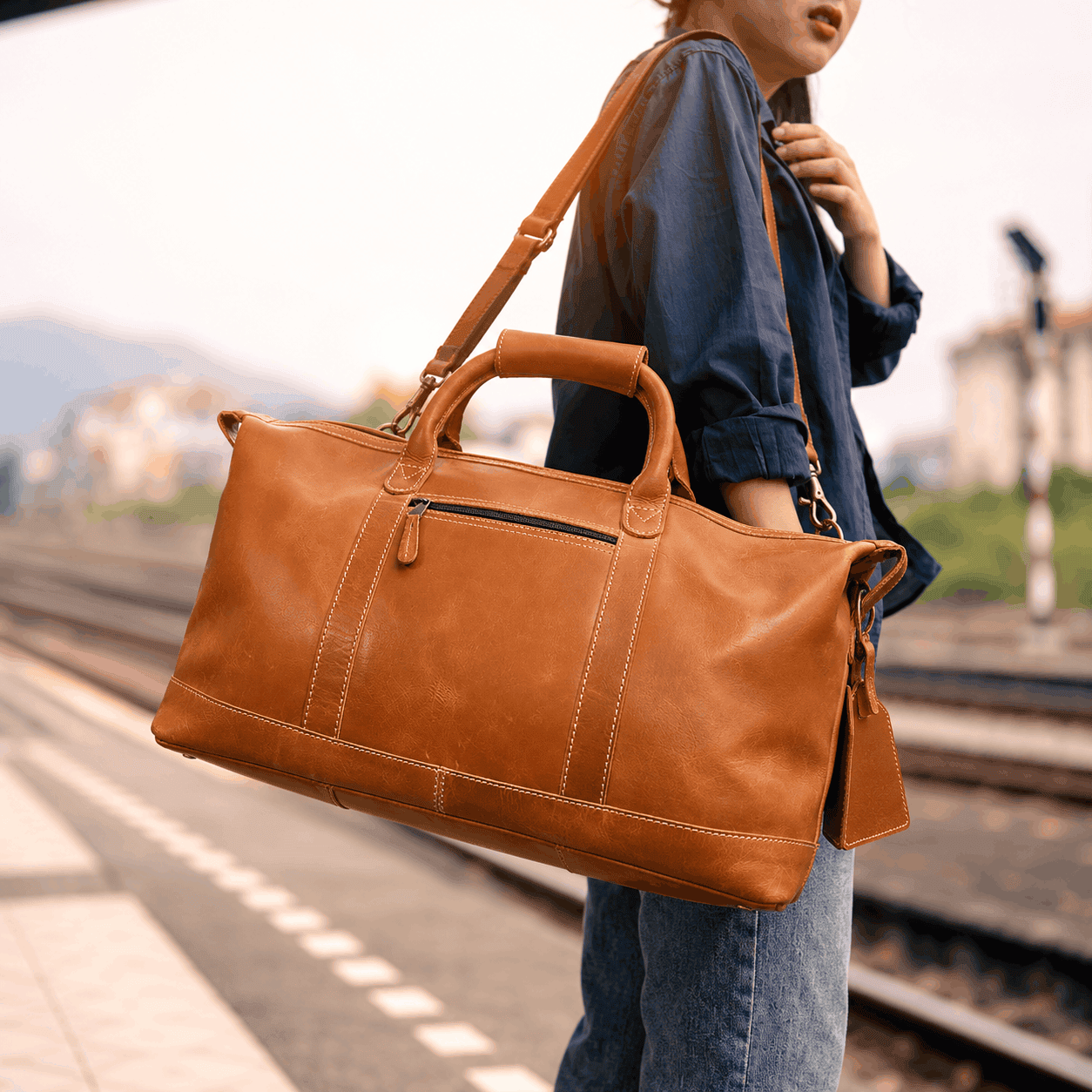 Cognac brown Boulder Canyon leather duffel bag with sturdy handles, adjustable shoulder strap, and front zippered pocket, carried by a woman at a train station.