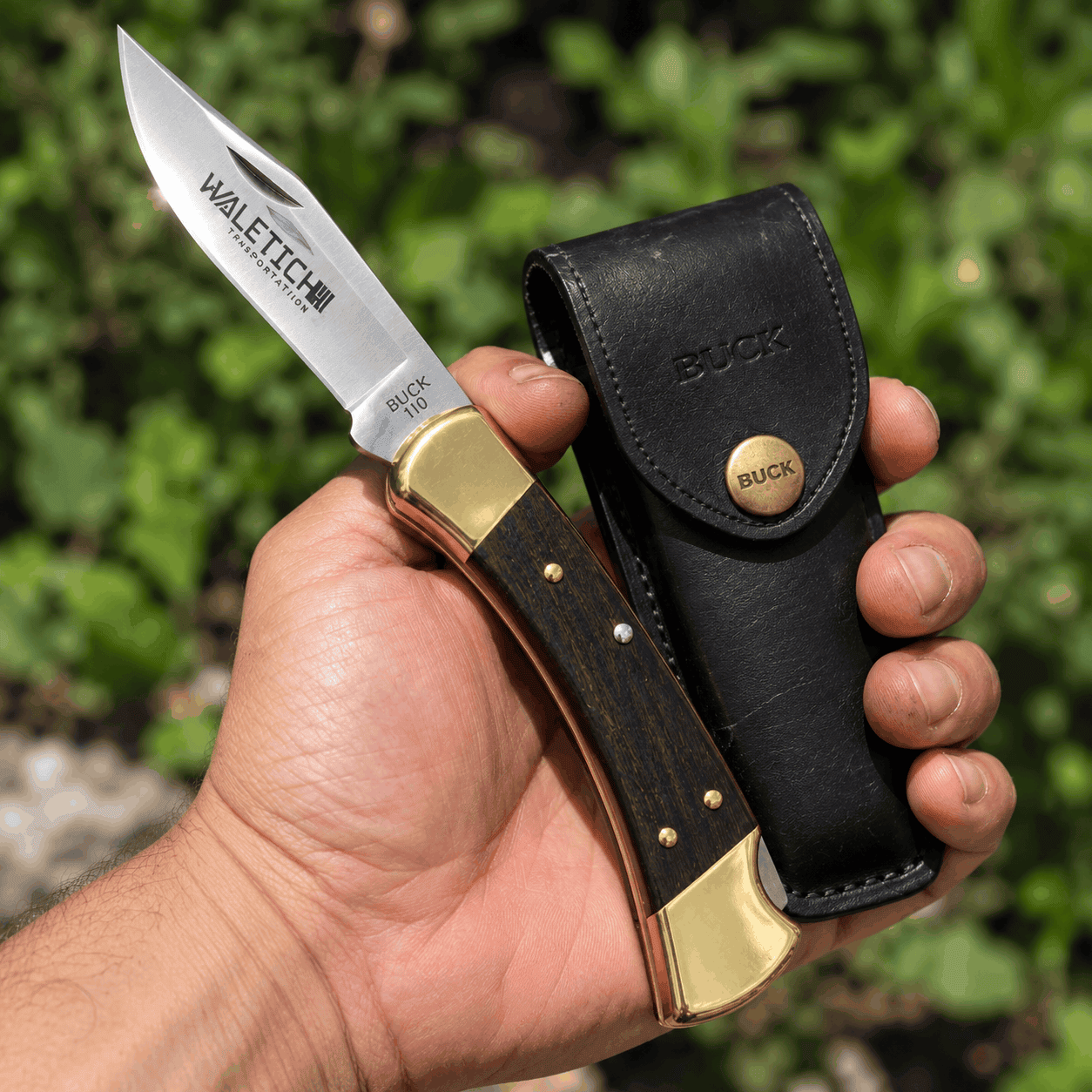 Buck 110 Folding Hunter knife with a wooden handle, brass accents, and custom engraved blade, held alongside its black leather sheath against a green outdoor background.