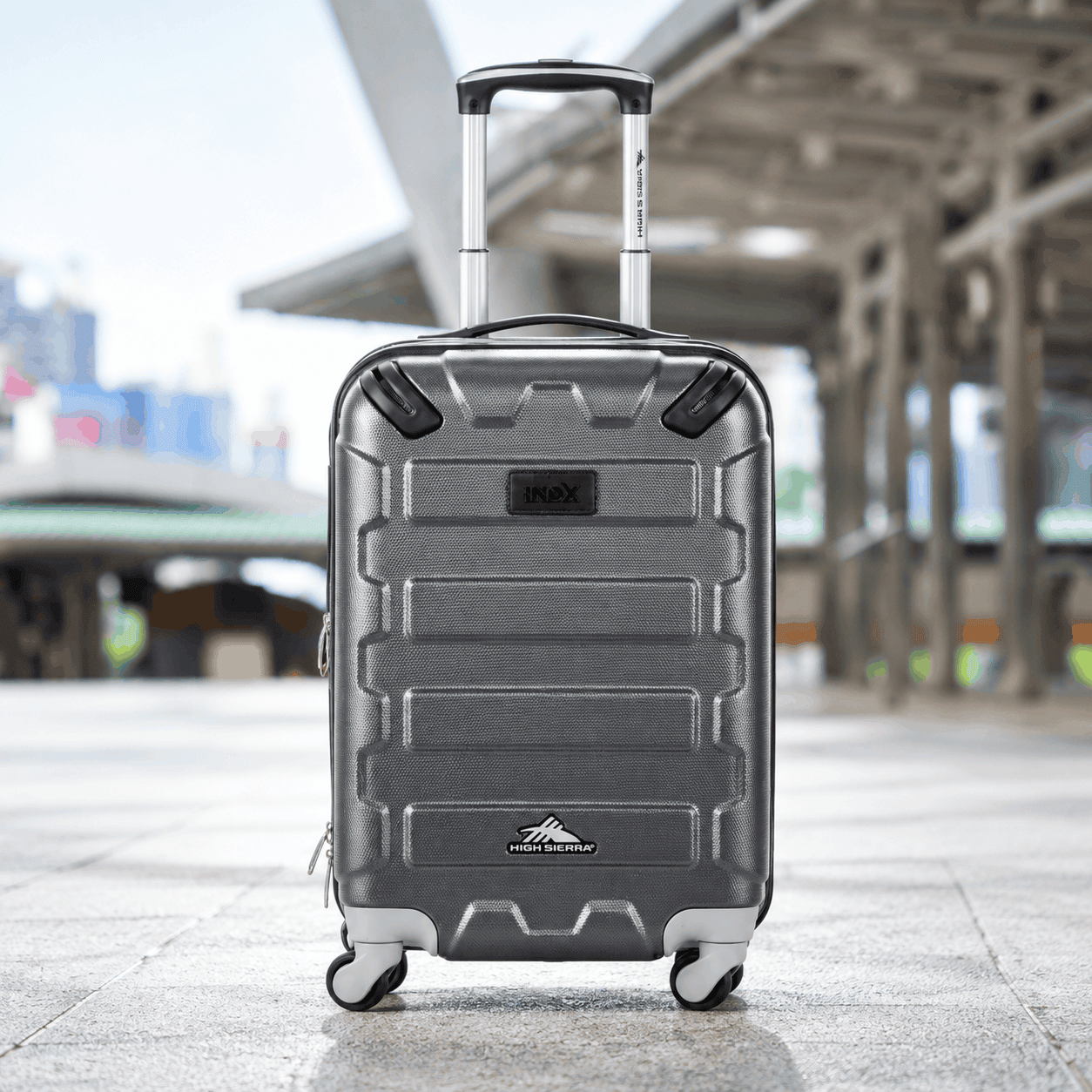 Gray High Sierra 20-inch hardside carry-on suitcase with a custom logo, textured protective shell, telescoping handle, and four spinner wheels at a train station.
