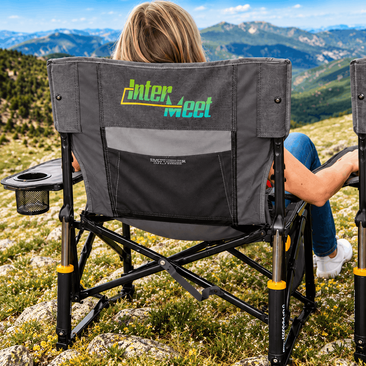 Gray GCI Outdoor Freestyle Rocker XL camping chair with a custom Intern Meet logo, built-in cup holder, and side table, overlooking a scenic mountain landscape.