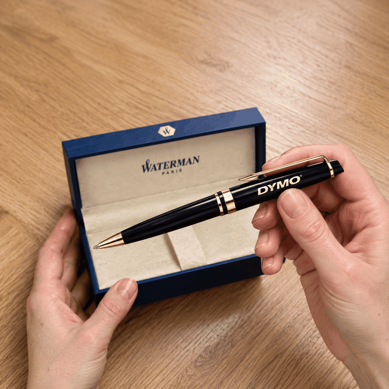 Black Waterman Expert ballpoint pen with gold trim and a custom DYMO logo, held above an open blue Waterman presentation gift box on a wooden tabletop.