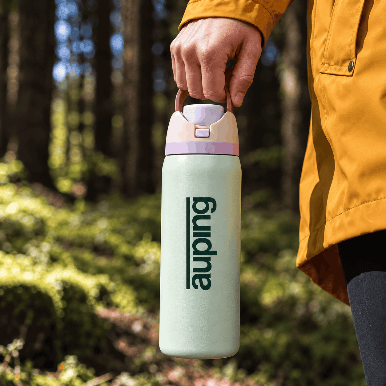 Light sage green Owala FreeSip 32 oz. water bottle with a lavender lid, built-in carrying handle, and custom black imprint, held by a person wearing a yellow jacket in a wooded setting.