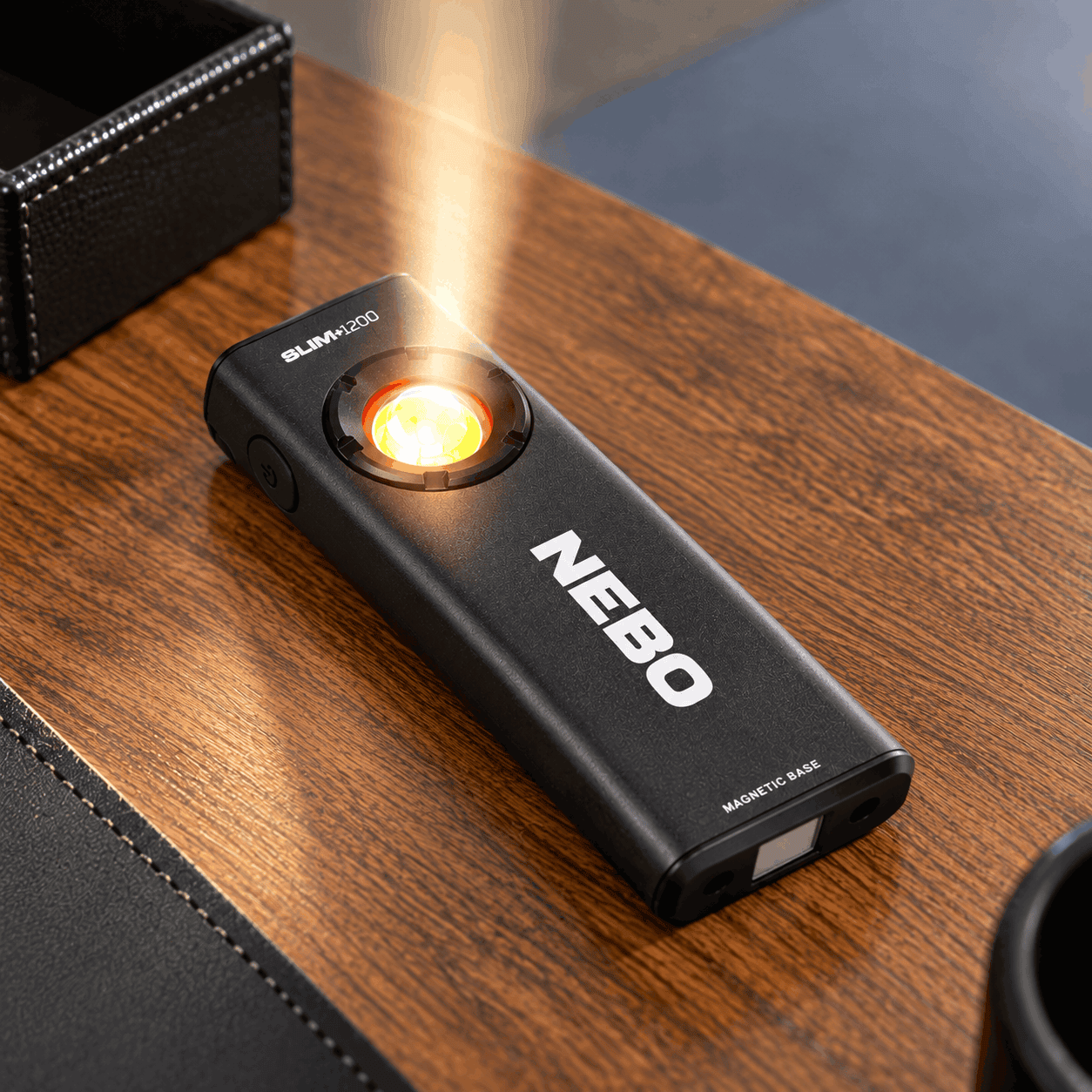 Black Nebo Slim+ 1200 rechargeable pocket light with a white NEBO logo and illuminated flashlight, resting on a wooden desk.