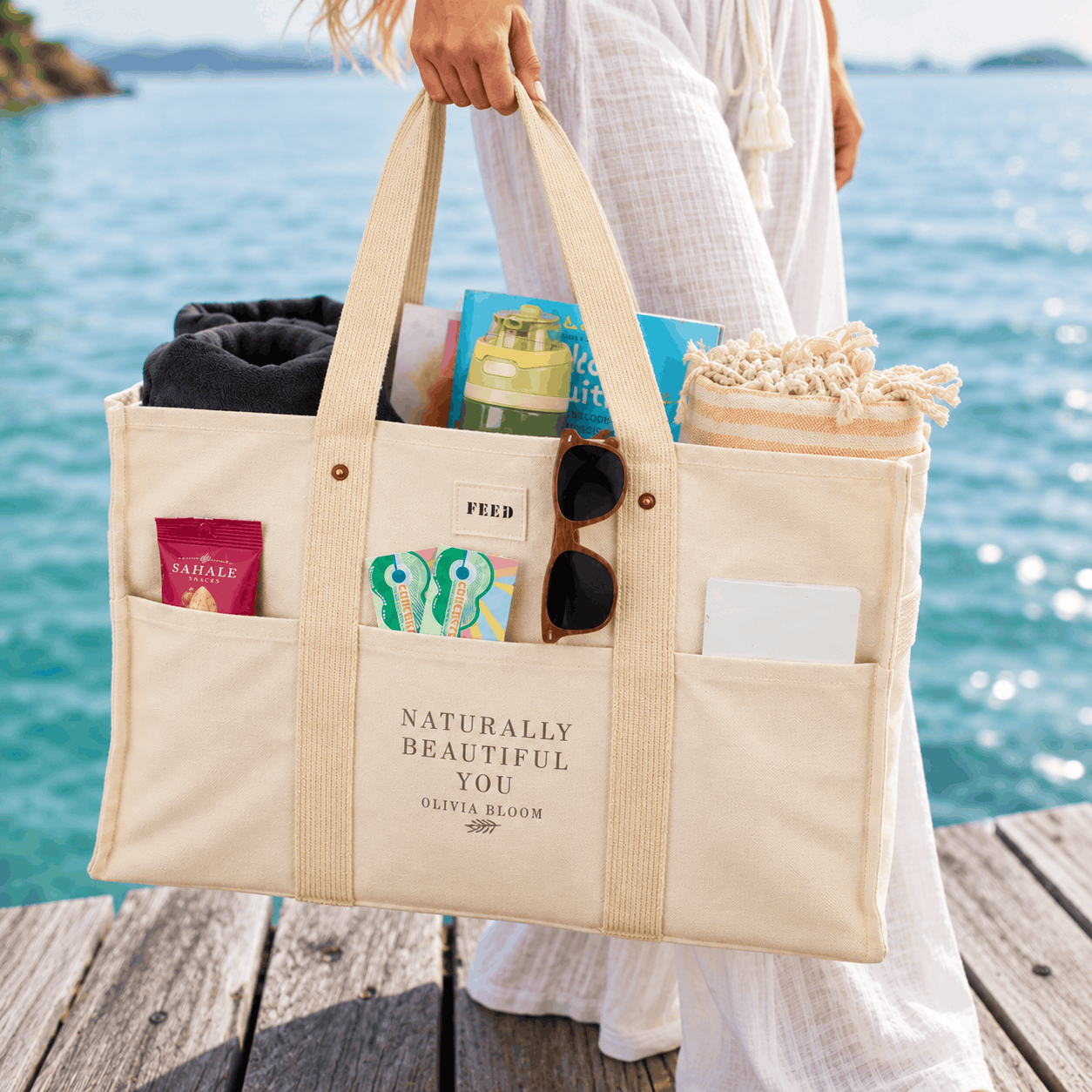 Natural cotton market tote bag with custom Olivia Bloom imprint, multiple exterior pockets, and beach essentials, carried by a woman beside the ocean.