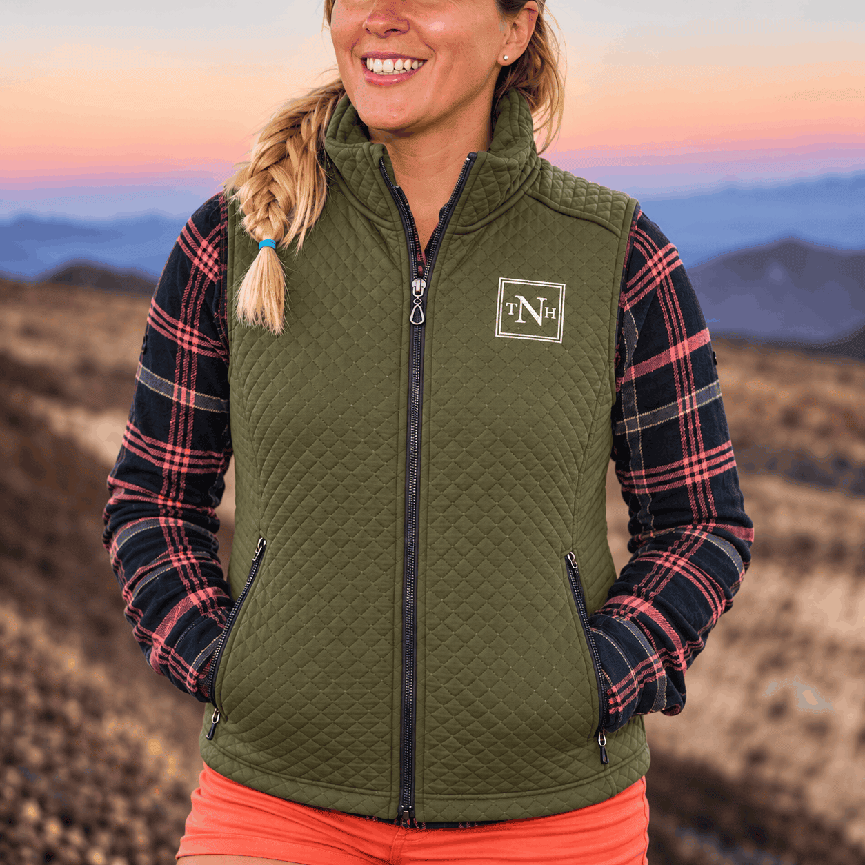 Olive green women's Mesa quilted vest with a custom embroidered TNH logo, full-zip front, and zippered pockets, worn over a plaid shirt in a scenic mountain setting.