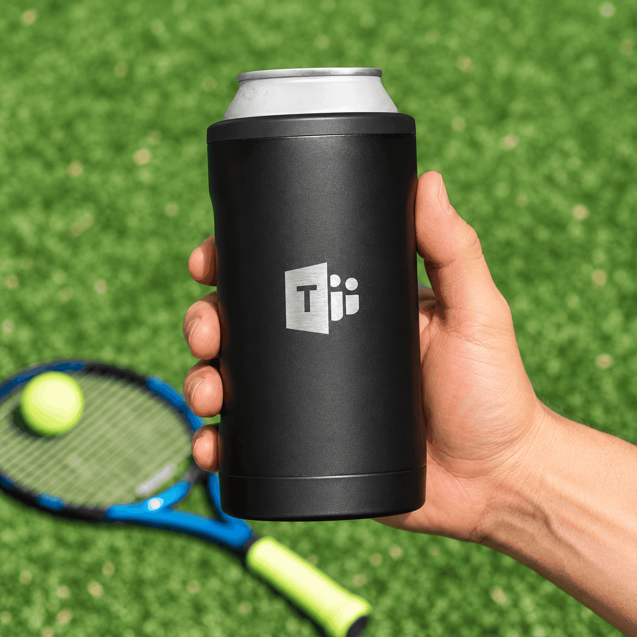 Black BrüMate Hopsulator TRiO 3-in-1 insulated can cooler with a custom silver logo, held over a grassy tennis court with a racket and tennis ball in the background.