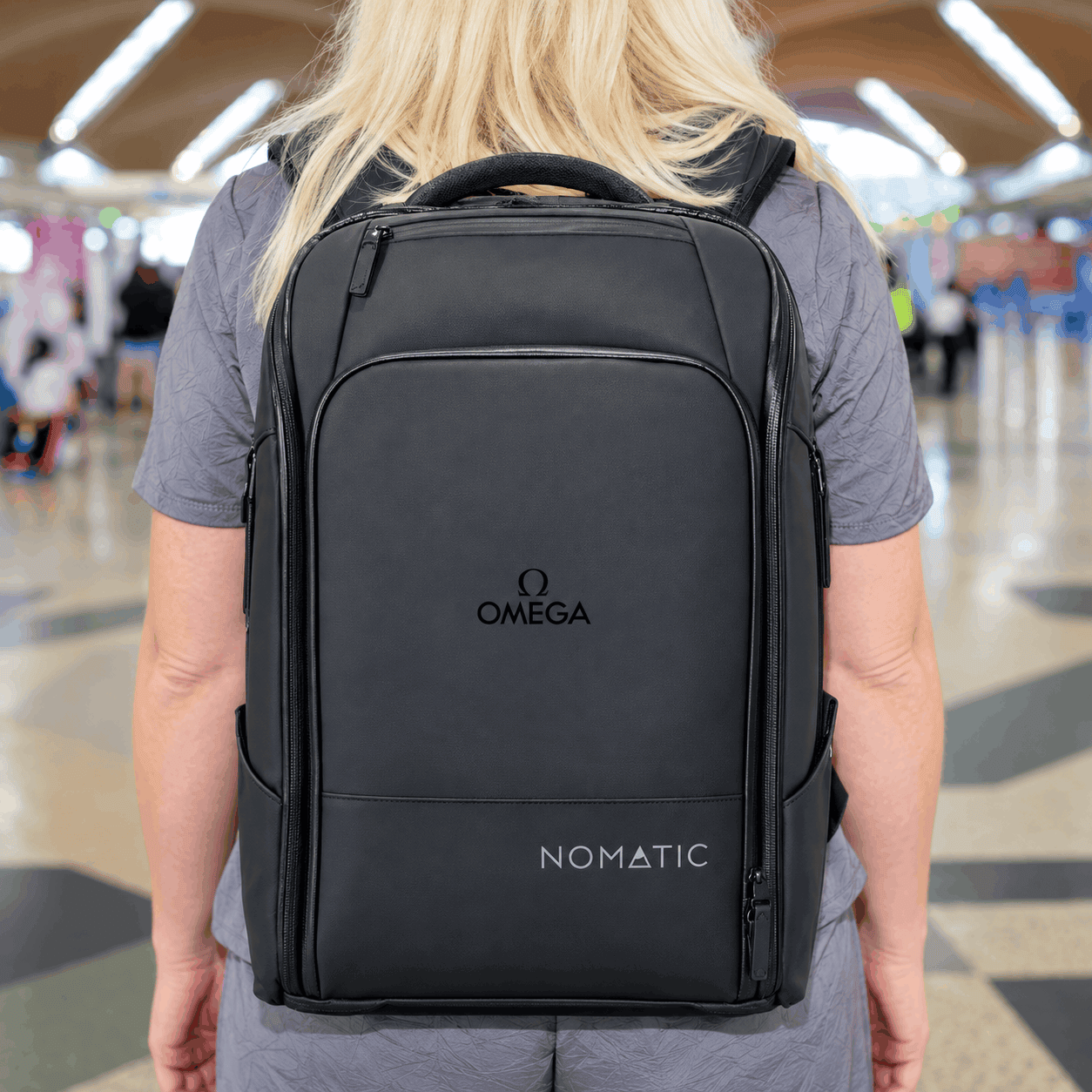 Black Nomatic 20L travel backpack with a custom Omega logo, multiple zippered compartments, and a sleek structured design, worn by a woman walking through an airport.