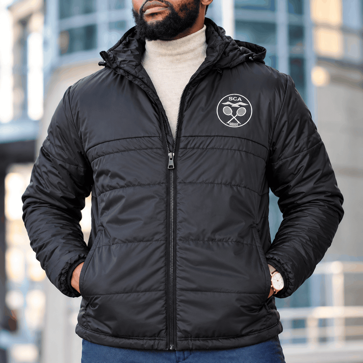 Black K2 quilted puffer jacket with a custom white embroidered chest logo, insulated hood, full-zip front, and side pockets, worn by a man in an urban setting.